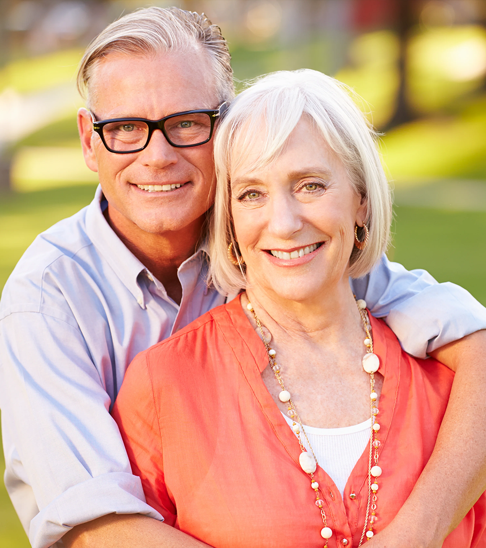 An older couple smiling, the man is hugging the women from behind.