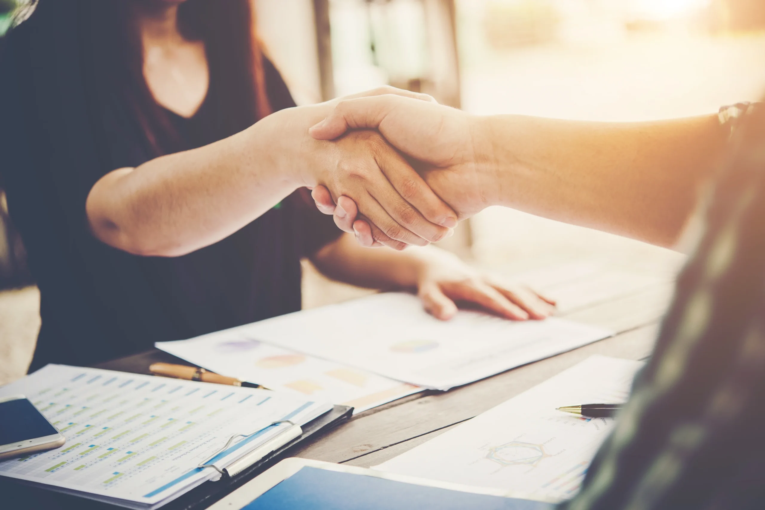 Two people shaking hands over a desk full of paperwork.